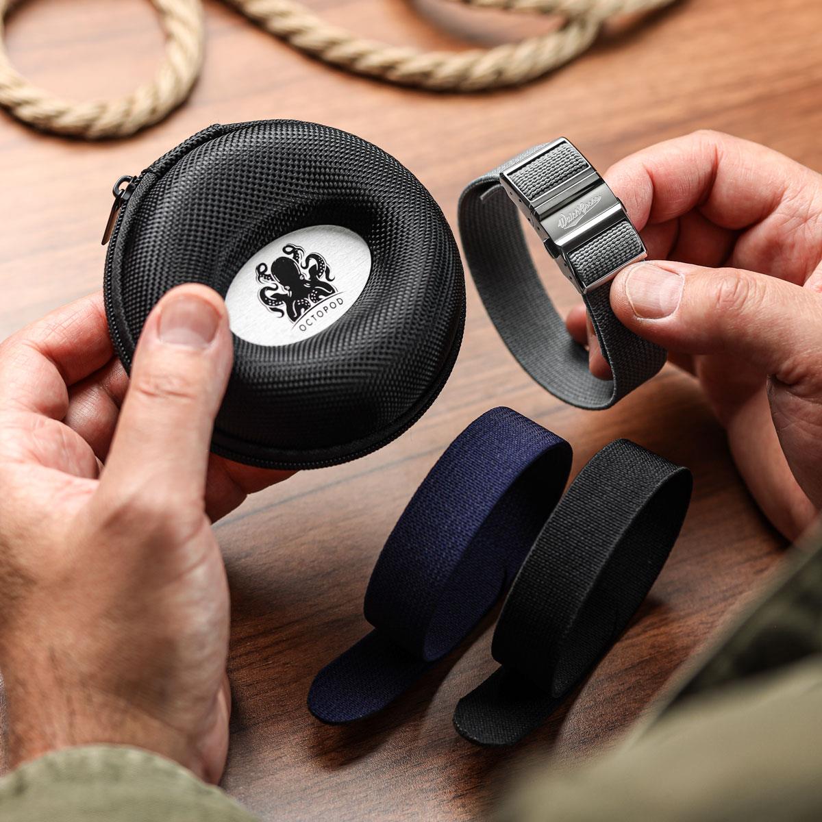 Person holding a black zippered case with a logo, surrounded by various straps on a wooden surface.