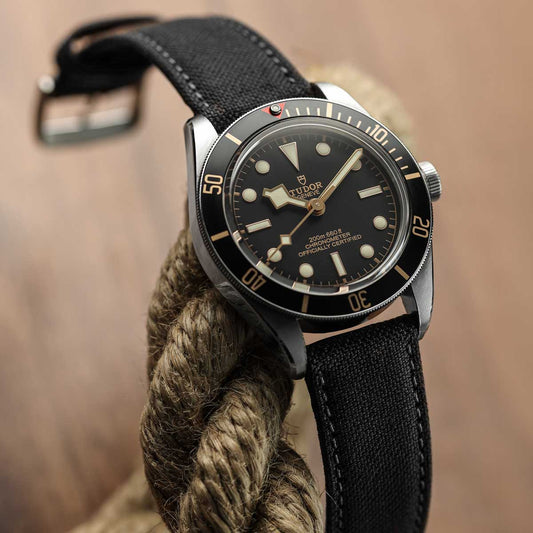 A close-up of a Tudor watch with a black dial, black bezel, and black fabric strap, resting on a thick brown rope against a warm brown background.