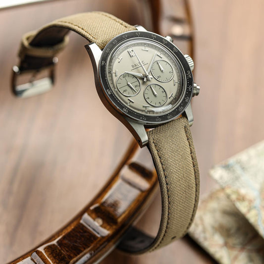 A vintage-style wristwatch with a beige canvas strap, a silver-toned metal case, and a light beige dial with three subdials. The watch is photographed resting on a brown leather strap against a warm wooden background.