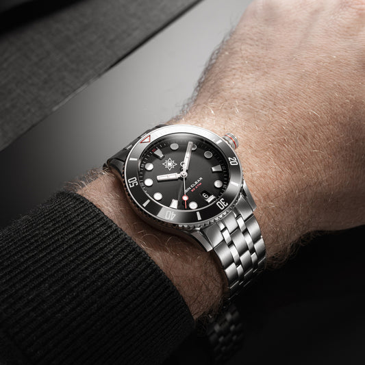 Close-up of a wrist wearing a black Second Hour Gin Clear wristwatch with a silver band on a dark background