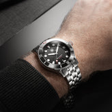 Close-up of a wrist wearing a black Second Hour Gin Clear wristwatch with a silver band on a dark background