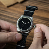 A close-up of a wristwatch with a black dial and black woven nylon strap, being adjusted by two hands over a wooden surface. Boldr Venture Umbra