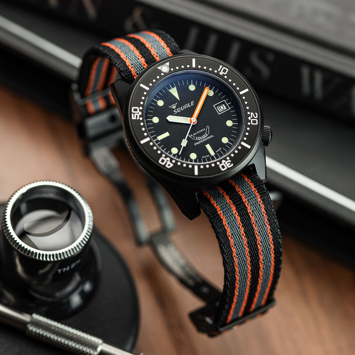 A wristwatch with a black dial, black bezel, and a woven black-and-orange strap, photographed on a wooden surface with a magnifying glass and books in the background. WatchGecko OCTOPOD Titanium MK3 Buckle - Super Black