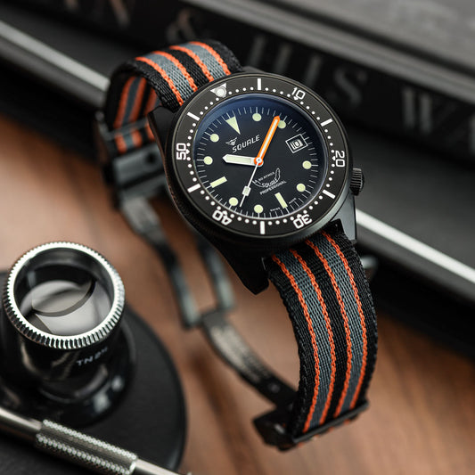 A wristwatch with a black dial, black bezel, and a woven black-and-orange strap, photographed on a wooden surface with a magnifying glass and books in the background. WatchGecko OCTOPOD Titanium MK3 Buckle - Super Black
