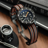 A wristwatch with a black dial, black bezel, and a woven black-and-orange strap, photographed on a wooden surface with a magnifying glass and books in the background. WatchGecko OCTOPOD Titanium MK3 Buckle - Super Black